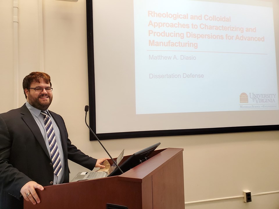 A man (me) in a suit is standing behind a podium near the left edge of the image. On the right is a projected screen showing a PowerPoint presentation title slide, with the title Rheological and Colloidal Approaches to Characterizing and Producing Dispersions for Advanced Manufacturing